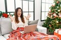 Beautiful caucasian woman using laptop sitting on the sofa by christmas tree crazy and mad shouting and yelling with aggressive Royalty Free Stock Photo