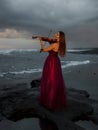 Beautiful Caucasian woman with violin on the beach. Music and art concept. Slim girl wearing long red dress and playing violin in Royalty Free Stock Photo