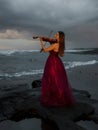 Beautiful Caucasian woman with violin on the beach. Music and art concept. Slim girl wearing long red dress and playing violin in Royalty Free Stock Photo