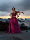 Beautiful Caucasian woman with violin on the beach. Music and art concept. Slim girl wearing long red dress, sitting on the rock Royalty Free Stock Photo