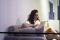 Beautiful Caucasian young woman with long dark hair sitting on sofa in modern living room typing on tablet browsing