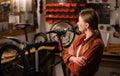 Beautiful Caucasian young woman repairing bicycle, looking on it with arm crossed. Bike workshop interior. Royalty Free Stock Photo