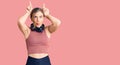 Beautiful caucasian young woman wearing gym clothes and using headphones doing funny gesture with finger over head as bull horns