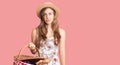 Beautiful caucasian young woman wearing summer hat and holding picnic wicker basket with bread thinking attitude and sober