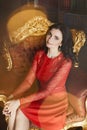 Beautiful and charming brunette model girl with curly hair in red stylish dress sits on the gold vintage armchair and Royalty Free Stock Photo