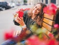 Beautiful charming young funny happy girl with curly hair and a smartphone in her hands sits on a bench in the city Royalty Free Stock Photo