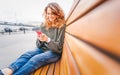 Beautiful charming young funny happy girl with curly hair and a smartphone in her hands sits on a bench in the city Royalty Free Stock Photo