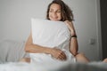 A beautiful charming young woman sits in bed and covers her body with a pillow, laughs joyfully and enjoys the new day Royalty Free Stock Photo