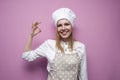 Beautiful cheerful girl cook in uniform and apron on a colored pink background shows okay, portrait of a housewife woman in