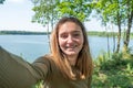 Beautiful cheerful young woman having a good time at the forest lakeside on a lovely day, taking a selfie, smiling