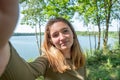 Beautiful cheerful young woman having a good time at the forest lakeside on a lovely day, taking a selfie, smiling