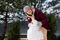 Beautiful cheery young woman in earflaps hat makes a snowman, having fun in a snow covered nature. Winter leisures