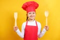 Beautiful chef child holds in his hands a wooden spoon and a spatula. Little girl in a cap and apron on a yellow background