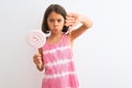 Beautiful child girl eating sweet lollipop standing over isolated white background with angry face, negative sign showing dislike