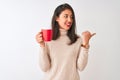 Beautiful chinese woman drinking red cup of coffee standing over isolated white background pointing and showing with thumb up to Royalty Free Stock Photo
