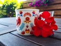 A beautiful coffee cup in hand-painted handmade folk patterns; red blooming geranium flower next to the cup set on a wooden table. Royalty Free Stock Photo