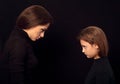 Beautiful concentrated angry mother and her seroius emotional daughter standing and looking each other on black shadow background