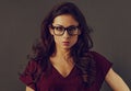 Beautiful concentrated serious brunette business woman looking in eye glasses with curly hair style in red silk blouse on grey