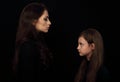 Beautiful concentrated strict mother and her serious emotional thinking daughter looking each other on black shadow background.