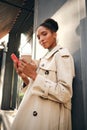 Beautiful confident African American girl in trench coat with coffee to go and cellphone sensually looking in camera Royalty Free Stock Photo
