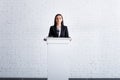 beautiful, confident lecturer standing on podium tribune near white brick wall.