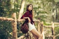 Beautiful country girl traveling and waiting near wooden fence wearing red checkered t-shirt, shorts and leather brown backpack