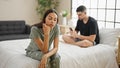 Beautiful couple disagreeing in their bedroom, sitting on the bed, expressing conflict while using a smartphone, a problem causing