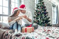 Beautiful couple in love, wearing santa hats, sitting next to a nicely decorated Christmas tree and making a toast with glasses of