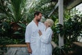 Beautiful couple in spa robes standing in hotel greenhouse, looking at each another, enjoying romantic wellness