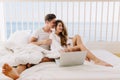 Beautiful couple spending time in home in summer morning, lying on bed with white laptop. Charming smiling girl with Royalty Free Stock Photo