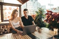 Beautiful couple in stylish casual clothes sitting in restaurant on terrace on sunset background and using laptop. Young man and Royalty Free Stock Photo