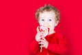 Beautiful curly baby girl eating a heart shaped candy on red background