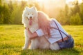 Beautiful curly blonde happy young woman in denim shorts hugging a white fluffy cute samoyed dog in the summer park Royalty Free Stock Photo