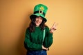 Beautiful curly hair woman wearing green hat with clover celebrating saint patricks day smiling with happy face winking at the