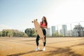 Beautiful curly mulatto girl in casual clothes stands with a longboard in hands on a tennis court, looks in camera and poses.