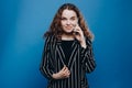 Beautiful curly young girl in casual wear looking at the camera speaks by phone while standing isolated over blue background Royalty Free Stock Photo