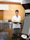 Male bakery worker wearing coat cap gloves holding bagel tray in bakery kitchen by prep table Royalty Free Stock Photo