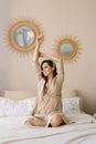 Beautiful cute brunette pregnant sitting on the bed in beige tones, raises her hands up, stretches and smiles Royalty Free Stock Photo