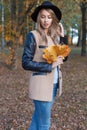 Beautiful cute cheerful girl in a black hat with a bouquet of bright colored leaves in a coat walks in autumn park Royalty Free Stock Photo