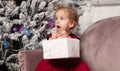 A beautiful cute little girl dressed in an elegant evening red dress sits on the couch and opens a New Year`s gift. Royalty Free Stock Photo