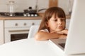 Beautiful cute little preschooler girl with dark hair sitting in front of laptop computer in kitchen, watching cartoons with Royalty Free Stock Photo