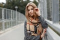 Beautiful cute young woman with blond hair in an elegant coat with a stylish beige scarf posing on a bridge in the city. Royalty Free Stock Photo