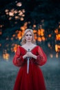 Beautiful, dangerous blonde queen in a red fashion lush dress with a dagger in her hands. Backdrop dark fantasy forest Royalty Free Stock Photo