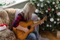 beautiful girl sits near the Christmas tree decorated with balls and light and playing the guitar and singing. Young Royalty Free Stock Photo