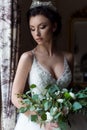 Beautiful delicate bride happy woman with a crown on her head by the window with a large wedding bouquet in a luxurious white