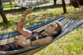 Stylish young woman with short dyed hair lying on a hammock in a sunny green park, listening to music and browsing internet on a Royalty Free Stock Photo