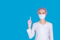 A beautiful doctor in a medical mask, cap and gloves shows a symbolic hand gesture-warning, attention, on a blue background.