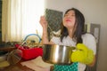 Lifestyle portrait of young pretty and happy Asian Chinese woman in apron holding cooking pot tasting delicious soup smiling