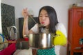 Lifestyle portrait of young pretty and happy Asian Korean woman in apron holding cooking pot tasting delicious soup smiling