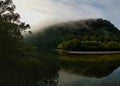 Beautiful early morning view of a creek with reflections of blue sky, foggy mountains and trees on water Royalty Free Stock Photo